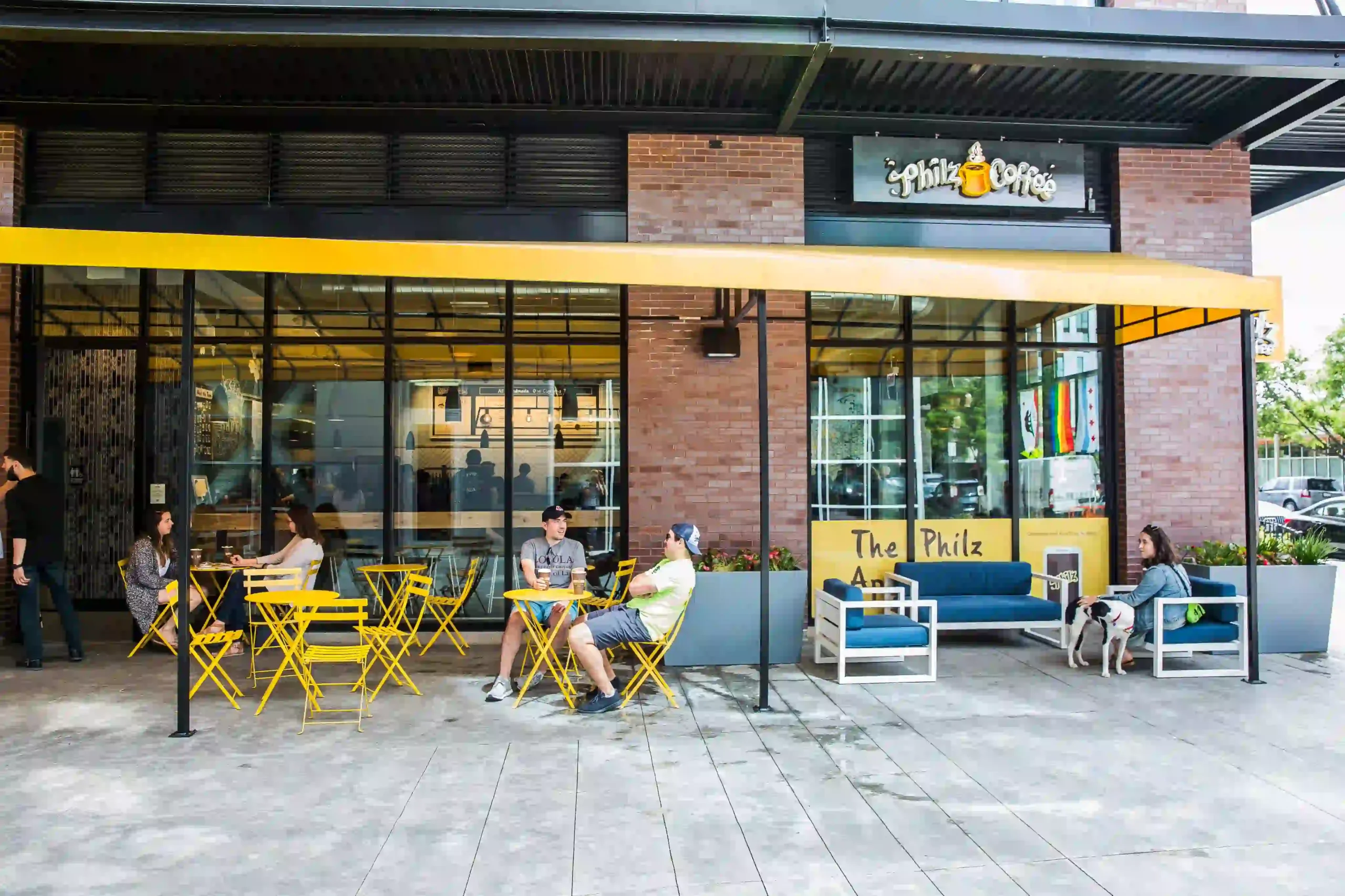 Philz Coffee storefront with large glass windows, a yellow awning, and outdoor tables and chairs; people enjoy drinks and relax on blue couches, with a dog also present in the seating area.