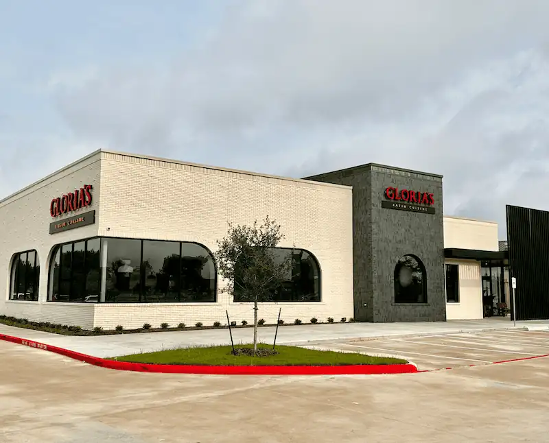 Exterior of Gloria’s Latin Cuisine with white and dark gray facade, large arched windows, signage above entrance, landscaped area with small tree, and parking lot in foreground.