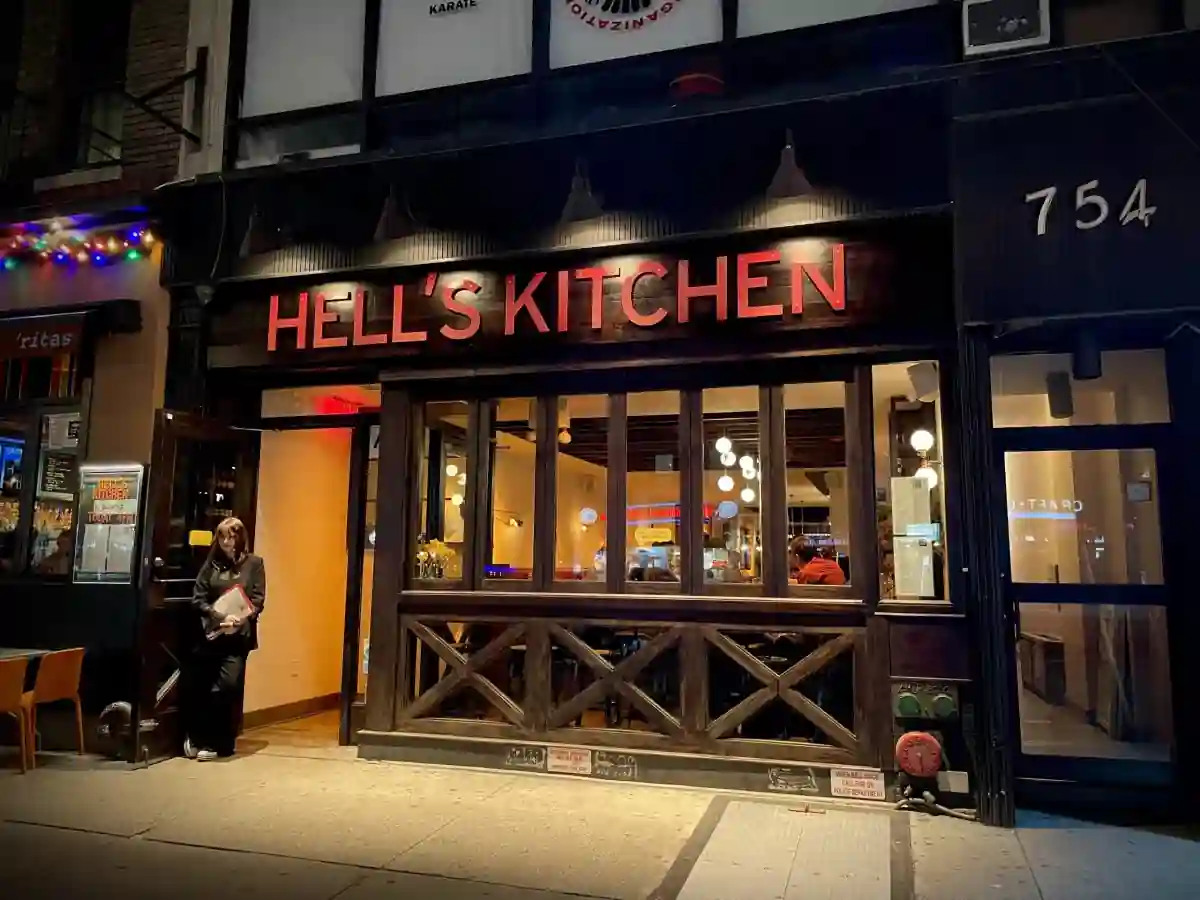 Nighttime street view of Hell's Kitchen restaurant exterior, showcasing its glowing red sign above the entrance, open doorway with visible indoor seating, and adjacent storefronts. The scene captures a cozy, urban dining atmosphere.