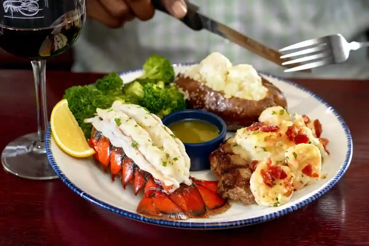 A plated surf and turf meal featuring lobster tail, steak topped with shrimp and bacon, baked potato with butter, and steamed broccoli. Served with a lemon wedge, melted butter dish, and a glass of red wine in a refined dining setting.