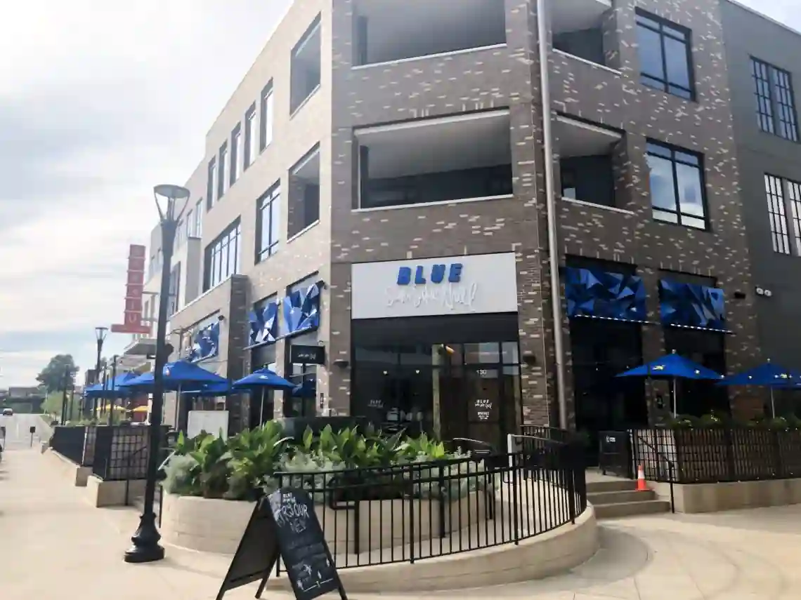 Street-level view of BLUE Sushi Sake Grill, a modern brick restaurant with large windows, outdoor seating with blue umbrellas, and a black metal fence along a tree-lined sidewalk.