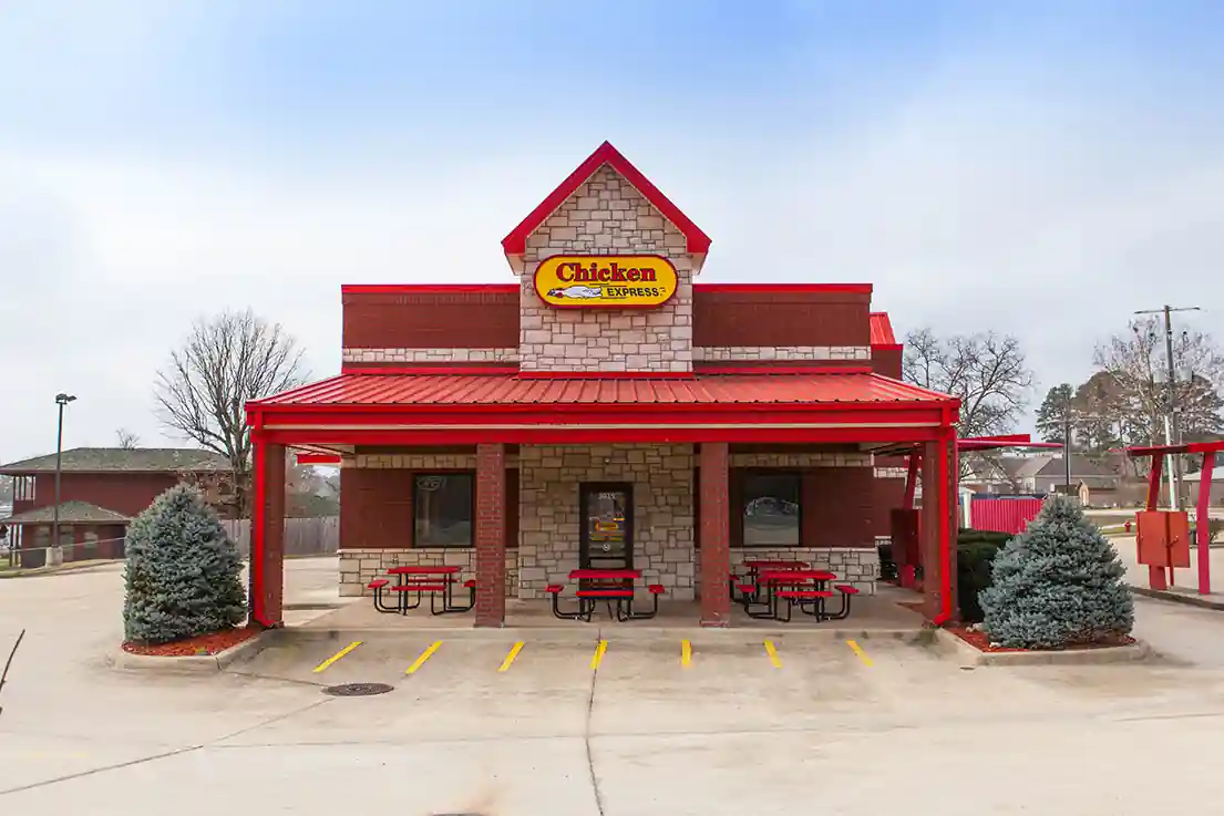 Exterior view of a Chicken Express restaurant with red and stone architecture, outdoor seating with red benches, and visible parking spaces.