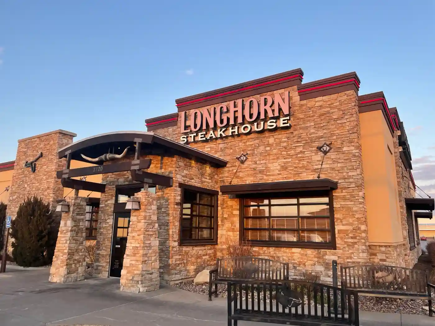 Exterior of LongHorn Steakhouse with rustic stone façade, “LONGHORN STEAKHOUSE” signage, mounted longhorn skull above entrance, address number “2720,” stone pillars, benches, and small landscaped area in foreground.