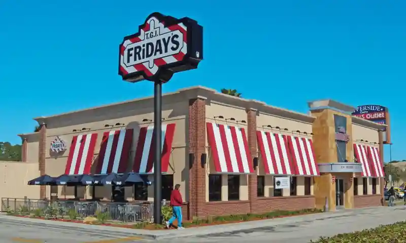 Image showing the front of T.G.I. Friday’s restaurant with striped awnings, a large branded sign, nearby Riverside Outlet, and a pedestrian walking by under a clear sunny sky.