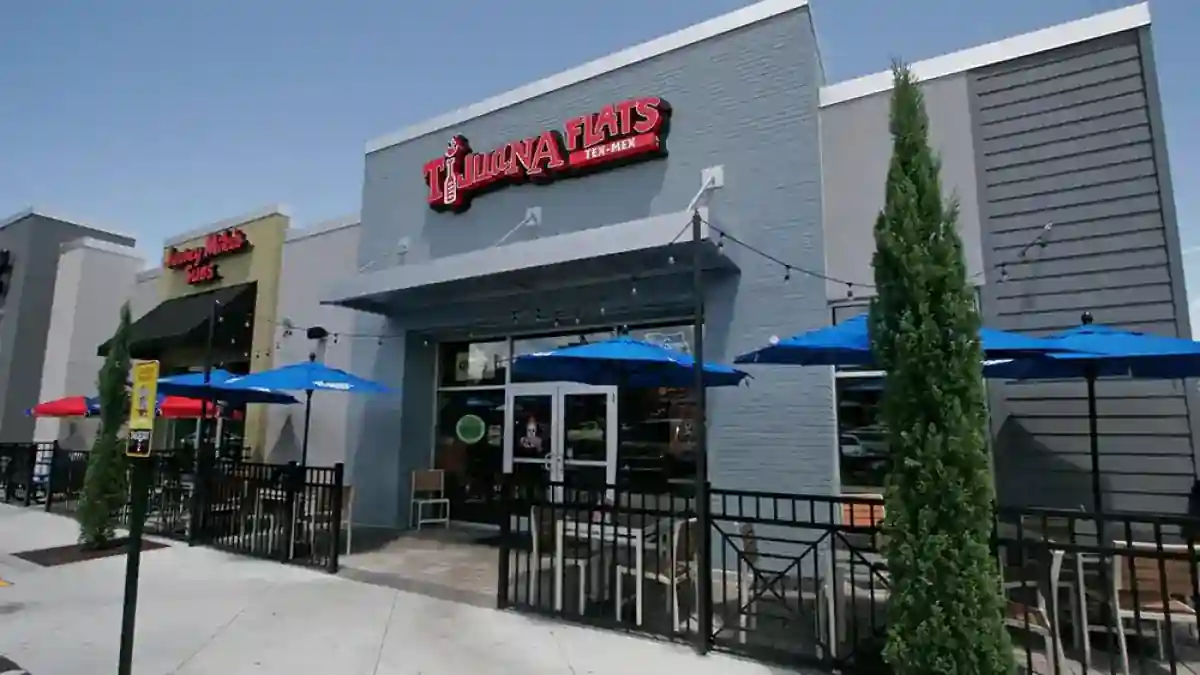 Exterior of Tijuana Flats and Jersey Mike’s Subs side by side, with colorful signage, fenced outdoor seating area shaded by red and blue umbrellas, and tall trees adding greenery in front of the patio.