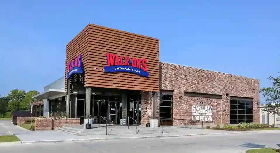 Exterior of Walk-On’s Bistreaux & Bar with red lettering on blue banner, large glass windows, wood-slatted awning, and painted mural stating “Game Day with a Taste of Louisiana.” Includes landscaped parking area.