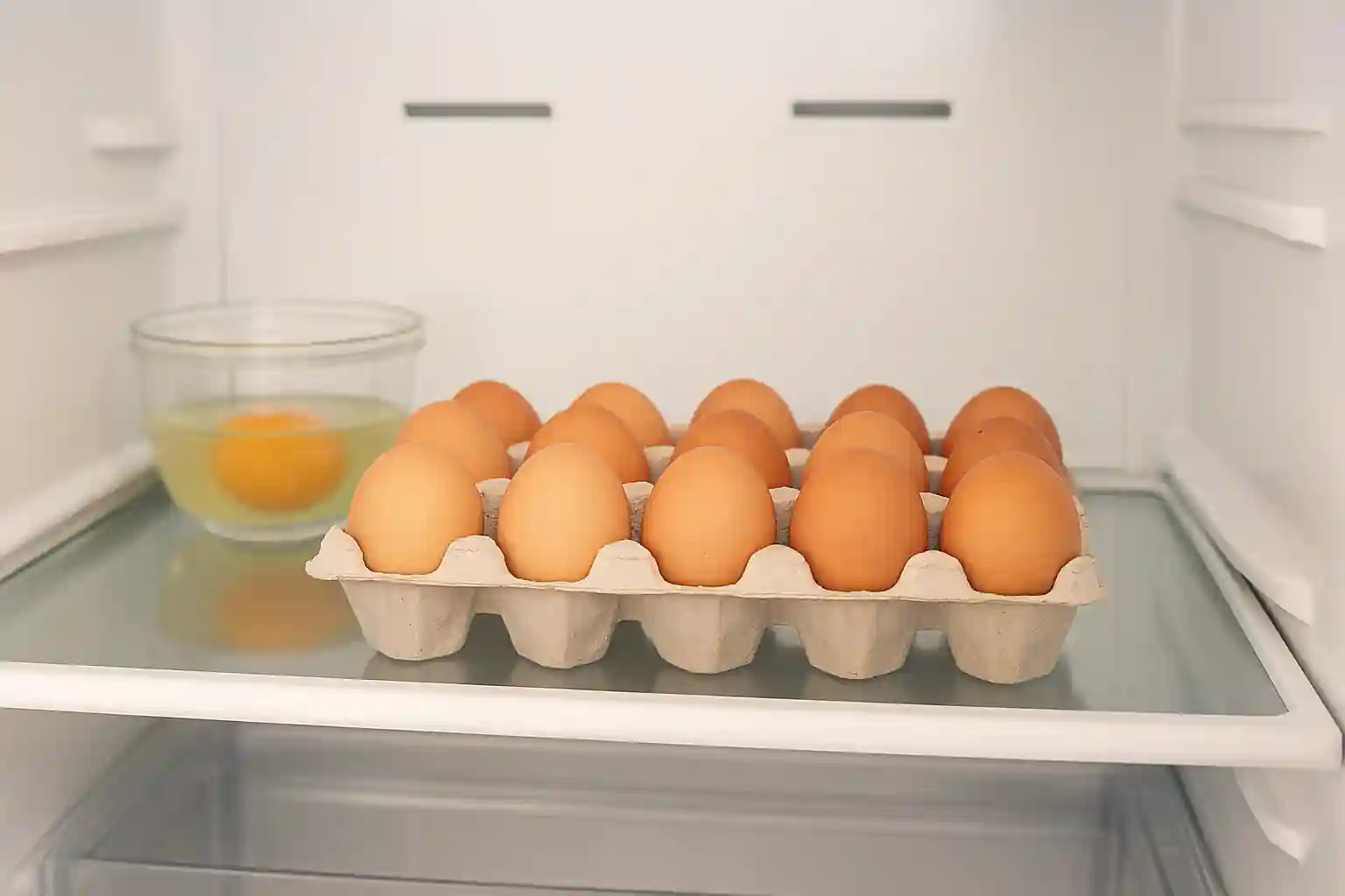 Carton of brown eggs stored on a refrigerator shelf with a cracked egg in a glass container nearby, illustrating proper egg storage techniques.