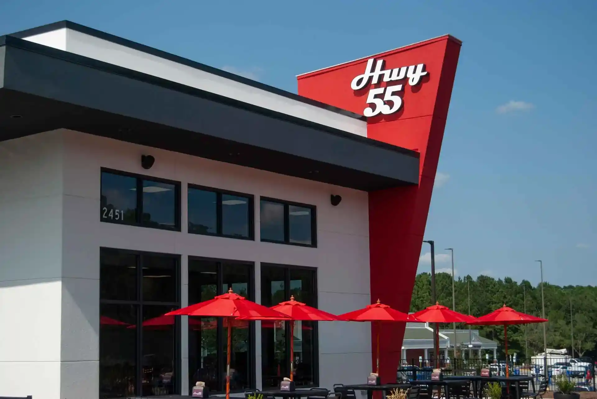 Exterior of Hwy 55 restaurant featuring red triangular sign with white text, white building with black trim and large windows, red patio umbrellas, and building number 2451.