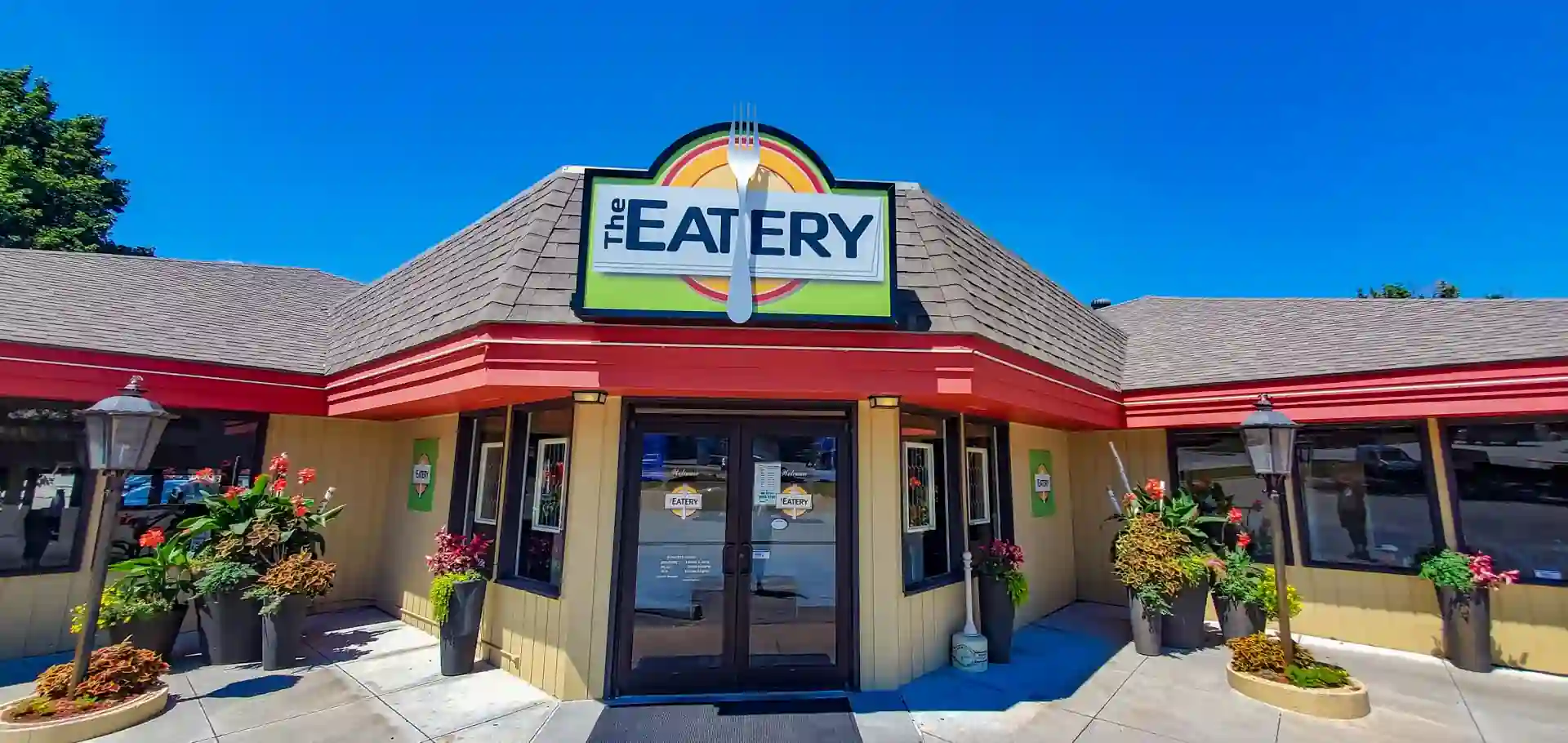 The Eatery restaurant exterior with beige and red facade, fork and spoon sign above entrance, double glass doors marked “Welcome,” flower pots on each side, and lamp posts in front under a clear blue sky.
