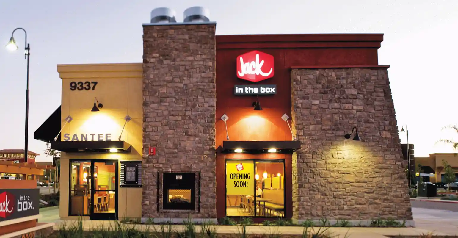 A Jack in the Box restaurant with a stone exterior, illuminated entrance, and a sign announcing its upcoming opening at 9337 Santee; interior visible through the glass doors.