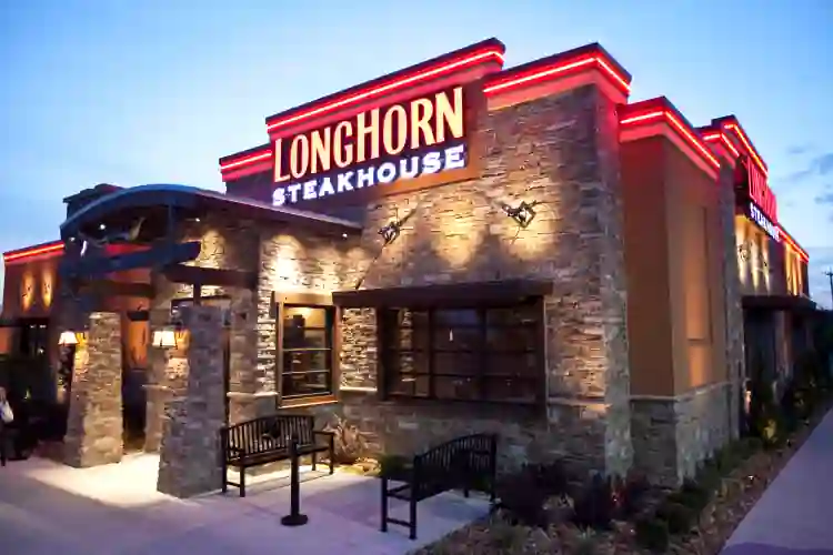 Exterior of LongHorn Steakhouse with illuminated red and white signage, stone walls, black benches in front, and landscaped greenery near entrance—set in warm lighting ambiance.
